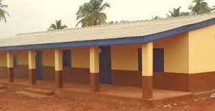 Single-story school building with a covered walkway, yellow walls, brown columns, and a metal roof on a dirt compound