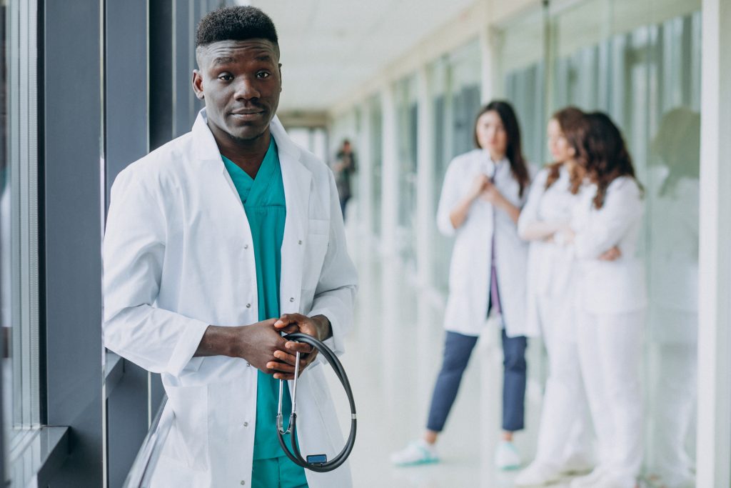 Healthcare professional in a white lab coat holding a stethoscope while standing in a hospital corridor, with colleagues in the background
