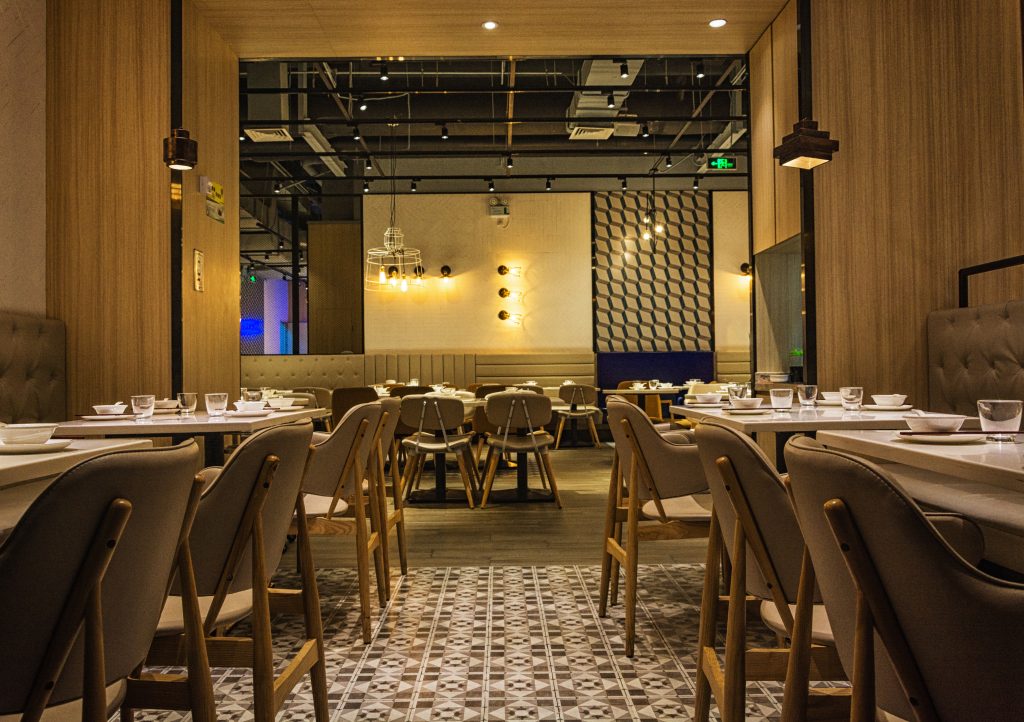 Modern restaurant dining room with neatly arranged tables and upholstered chairs, warm pendant lighting, wood-paneled walls, patterned tile flooring, and a contemporary, inviting atmosphere