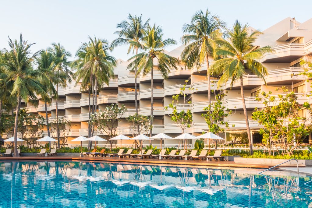 Large outdoor resort swimming pool surrounded by palm trees, lounge chairs, and white umbrellas, with a multi-story hotel building in the background